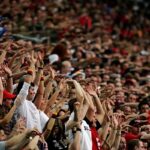 Large crowd at a stadium with arms raised, cheering fans likely at a sporting event in progress.