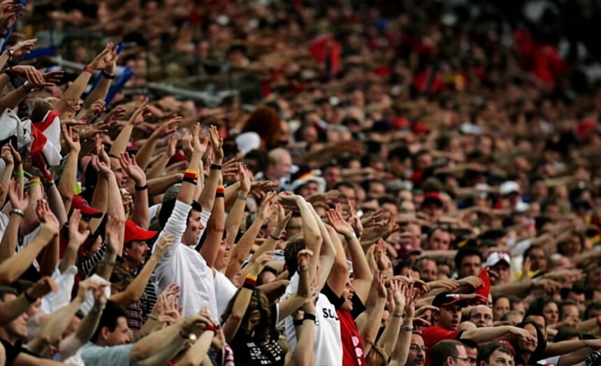 Large crowd at a stadium with arms raised, cheering fans likely at a sporting event in progress.