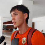 Young man in an orange shirt and backpack being interviewed by reporters in a bright indoor lobby.