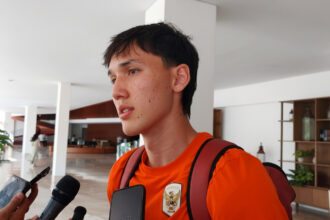 Young man in an orange shirt and backpack being interviewed by reporters in a bright indoor lobby.