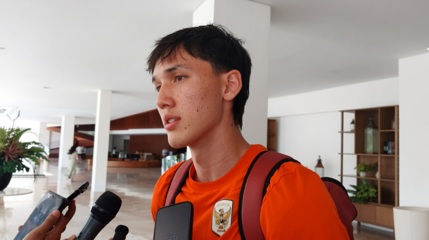 Young man in an orange shirt and backpack being interviewed by reporters in a bright indoor lobby.