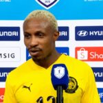 Football player in a yellow jersey speaks into a blue microphone during a post-match interview, backdrop covered with sponsor logos like Hyundai and Shopee.