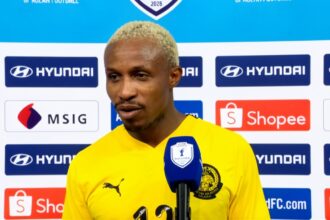 Football player in a yellow jersey speaks into a blue microphone during a post-match interview, backdrop covered with sponsor logos like Hyundai and Shopee.