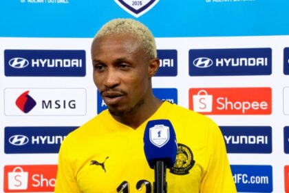 Football player in a yellow jersey speaks into a blue microphone during a post-match interview, backdrop covered with sponsor logos like Hyundai and Shopee.