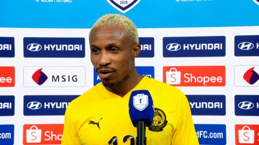 Football player in a yellow jersey speaks into a blue microphone during a post-match interview, backdrop covered with sponsor logos like Hyundai and Shopee.