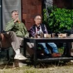 Four men sit around a low table outdoors, one speaking into a microphone, cups and snacks on the table, green plants in the background.