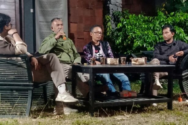 Four men sit around a low table outdoors, one speaking into a microphone, cups and snacks on the table, green plants in the background.