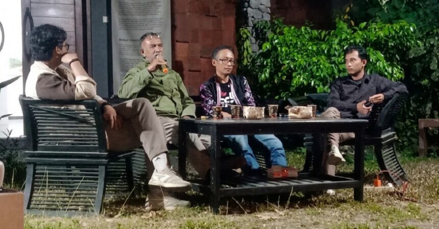 Four men sit around a low table outdoors, one speaking into a microphone, cups and snacks on the table, green plants in the background.