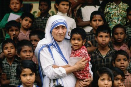 Older woman in a white and blue sari holding a young child, surrounded by curious children onlookers