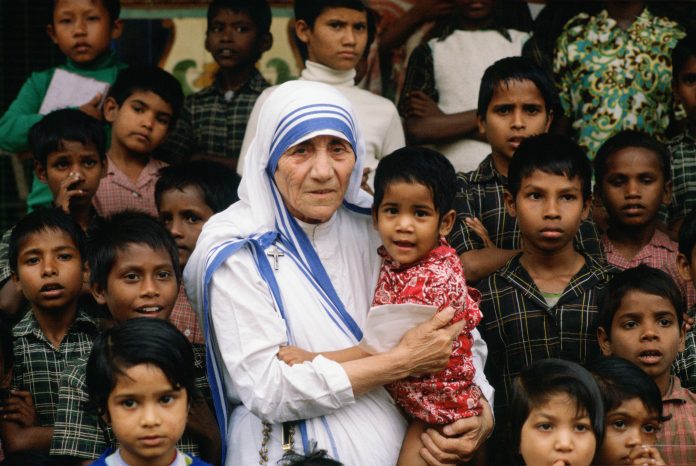 Older woman in a white and blue sari holding a young child, surrounded by curious children onlookers