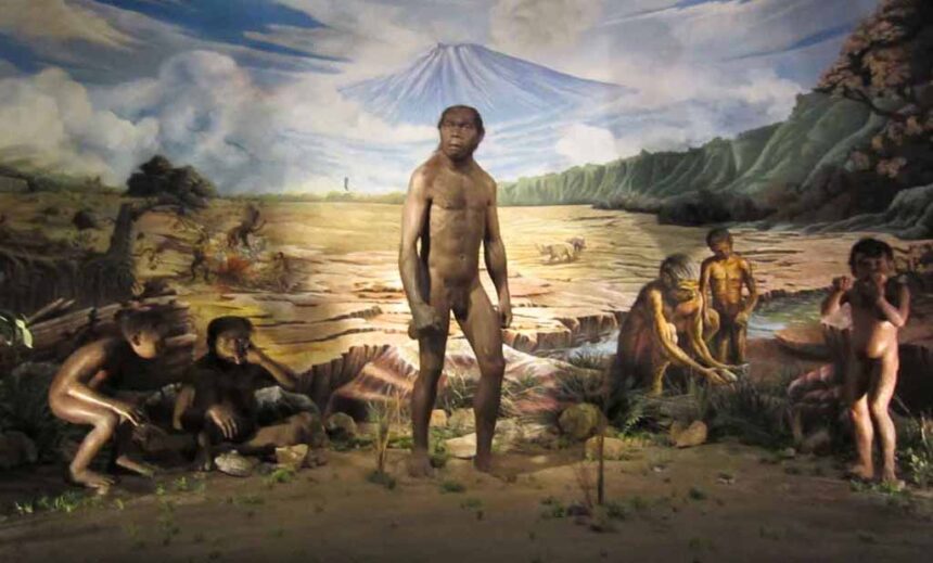 Life‑size prehistoric scene with nude human statues around a central adult figure in a barren landscape and a mural of mountains and a volcano in the background, suggesting an early human encampment.