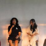 Four women panelists seated on stage during a discussion; one speaker holds a microphone while others listen.