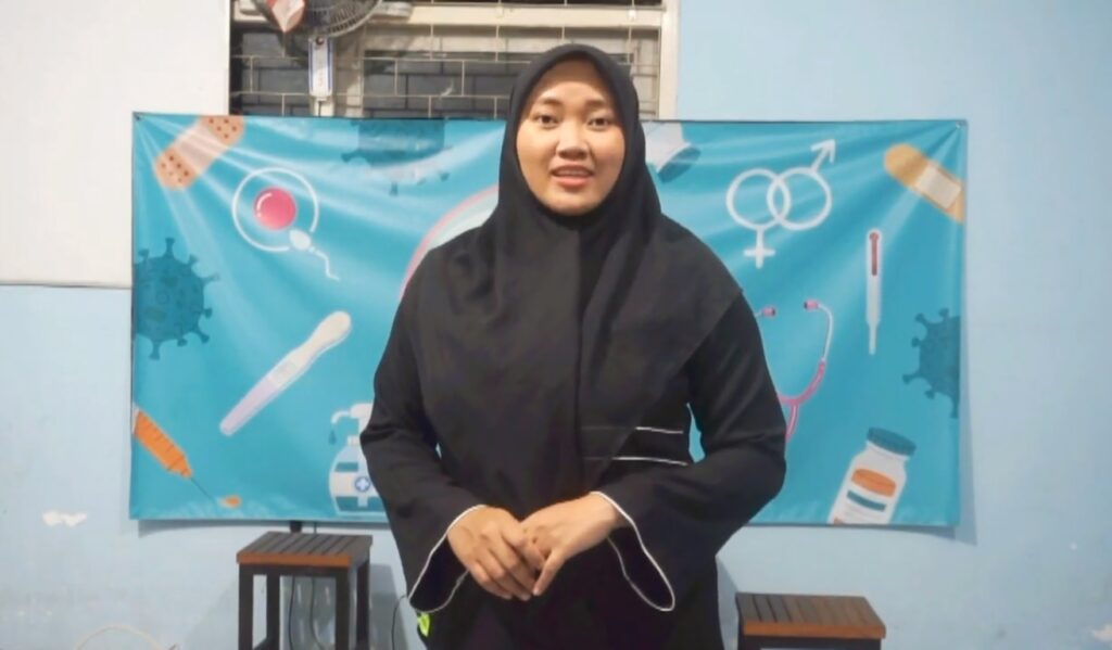Woman in a black hijab and robe speaks to an audience in front of a blue medical-themed backdrop.