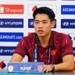 Vietnamese male footballer speaking at a press conference, seated at a table with a microphone and sponsor backdrop behind him.