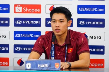 Vietnamese male footballer speaking at a press conference, seated at a table with a microphone and sponsor backdrop behind him.