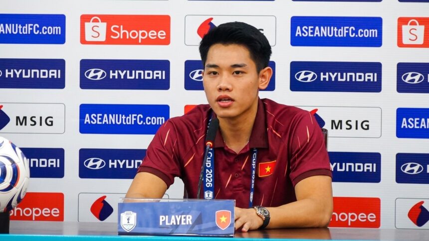 Vietnamese male footballer speaking at a press conference, seated at a table with a microphone and sponsor backdrop behind him.