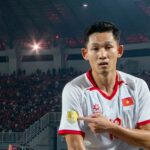 Vietnamese footballer in a white jersey with red trim points to a gold emblem on his sleeve amid a packed arena crowd