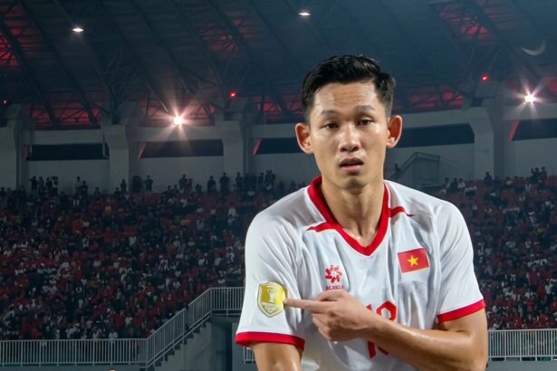 Vietnamese footballer in a white jersey with red trim points to a gold emblem on his sleeve amid a packed arena crowd