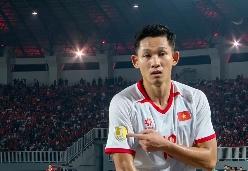 Vietnamese footballer in a white jersey with red trim points to a gold emblem on his sleeve amid a packed arena crowd