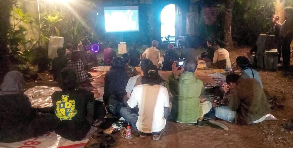 Outdoor night movie screening: a crowd sits on mats facing a projector screen in a garden setting.