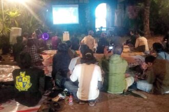 Outdoor night movie screening: a crowd sits on mats facing a projector screen in a garden setting.