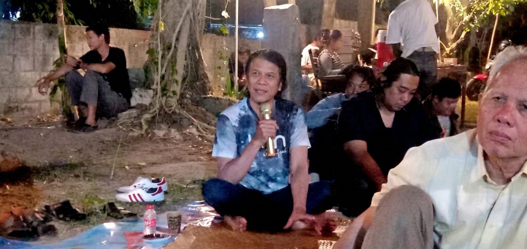 Man sitting cross-legged outdoors at night, holding a microphone and speaking to a small group.
