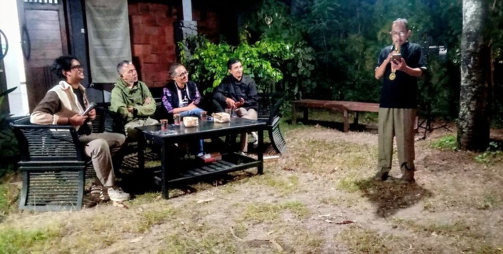 An outdoor evening gathering: five men seated around a table, one man standing and speaking into a microphone with a medal visible around his neck.