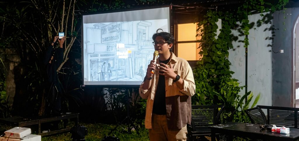 Man in a tan jacket speaks into a microphone at an outdoor presentation, with a sketched city scene on the projector behind him on screen.