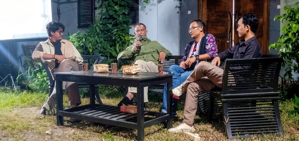 Four men sit on black outdoor furniture around a coffee table in a garden, one holding a microphone.