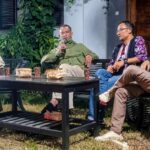 Four men sit on black outdoor furniture around a coffee table in a garden, one holding a microphone.