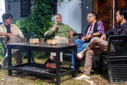 Four men sit on black outdoor furniture around a coffee table in a garden, one holding a microphone.