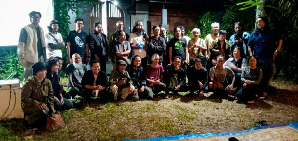 Group of about 25 people posing for a photo outdoors at night, arranged in two rows with some kneeling in front, casual clothing and greenery in the background.