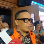 Man in an orange safety vest speaks to reporters, microphones in foreground and people listening nearby.