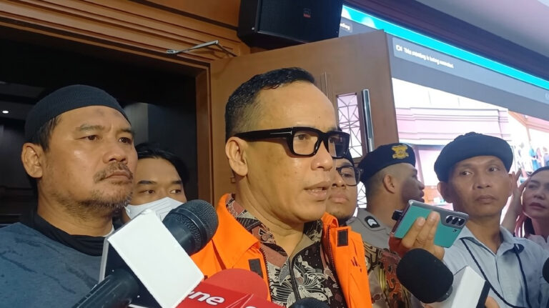 Noel Ebenezer by Kompas com reporter on location FIXX - Mabur.co Man in an orange safety vest speaks to reporters, microphones in foreground and people listening nearby.