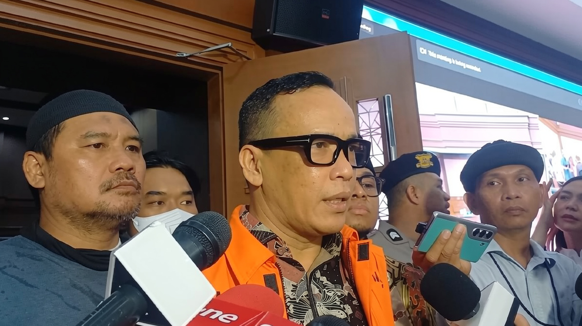 Man in an orange safety vest speaks to reporters, microphones in foreground and people listening nearby.