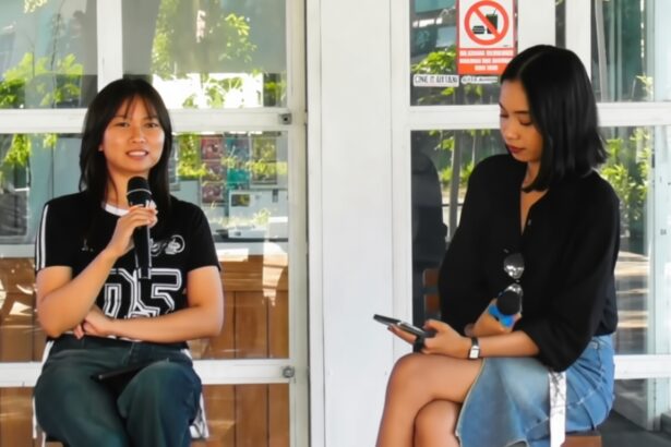 Two women sit on a bench outside a glass-walled building; the left woman speaks into a handheld microphone while the right woman looks at her smartphone and holds sunglasses.