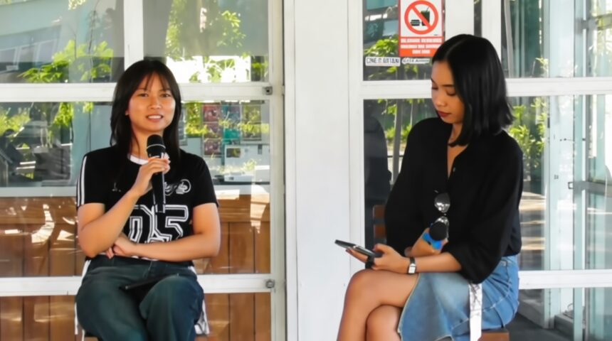Nulis Bareng Ananta FIXX - Mabur.co Two women sit on a bench outside a glass-walled building; the left woman speaks into a handheld microphone while the right woman looks at her smartphone and holds sunglasses.