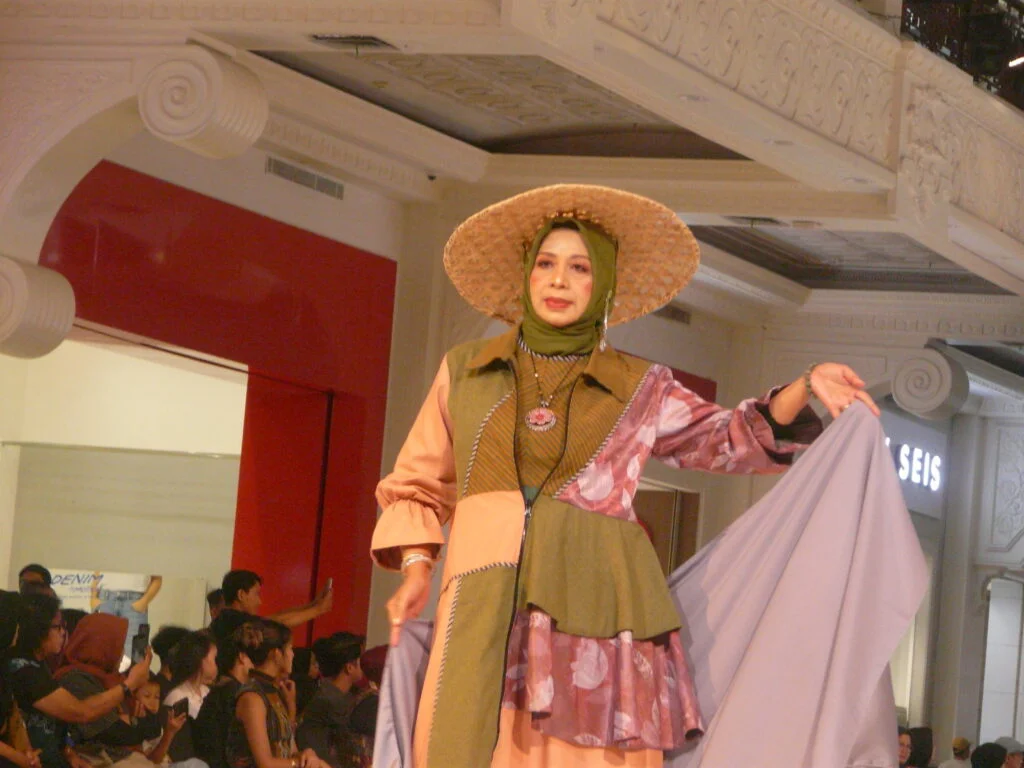 Model wearing a patchwork outfit and wide straw hat on a runway, holding a flowing lavender cape.