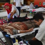 Donors rest on medical beds at a community blood donation camp as a nurse in a red hijab assists a patient nearby.