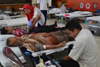 Donors rest on medical beds at a community blood donation camp as a nurse in a red hijab assists a patient nearby.