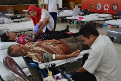 Donors rest on medical beds at a community blood donation camp as a nurse in a red hijab assists a patient nearby.