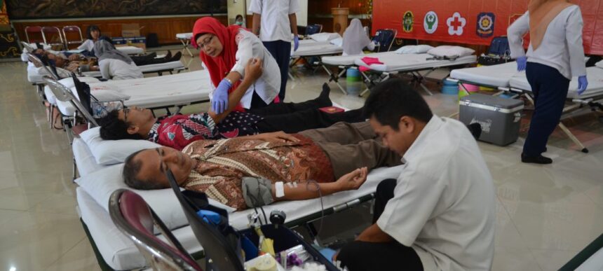Donors rest on medical beds at a community blood donation camp as a nurse in a red hijab assists a patient nearby.