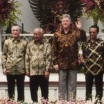Six men in patterned batik shirts standing on a stage with flowers, one man waving at the camera.