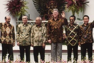 Six men in patterned batik shirts standing on a stage with flowers, one man waving at the camera.