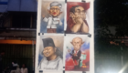 Outdoor display board with four caricature posters of politicians, taped to a white panel beside a fence and trees. The drawings exaggerate facial features and poses for humorous effect, arranged in a 2x2 grid.