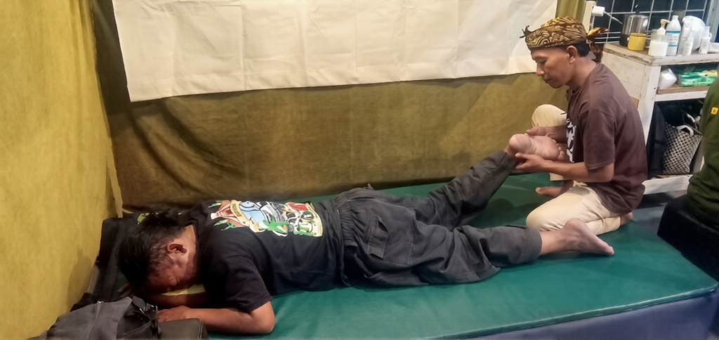 Man lying face down on a mat while another person holds and bends his leg in a training or therapy setting.