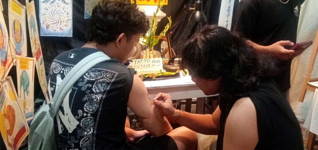 Tattoo artist working on a man's upper arm in a shop, with posters and equipment around.