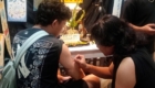 Tattoo artist working on a man's upper arm in a shop, with posters and equipment around.