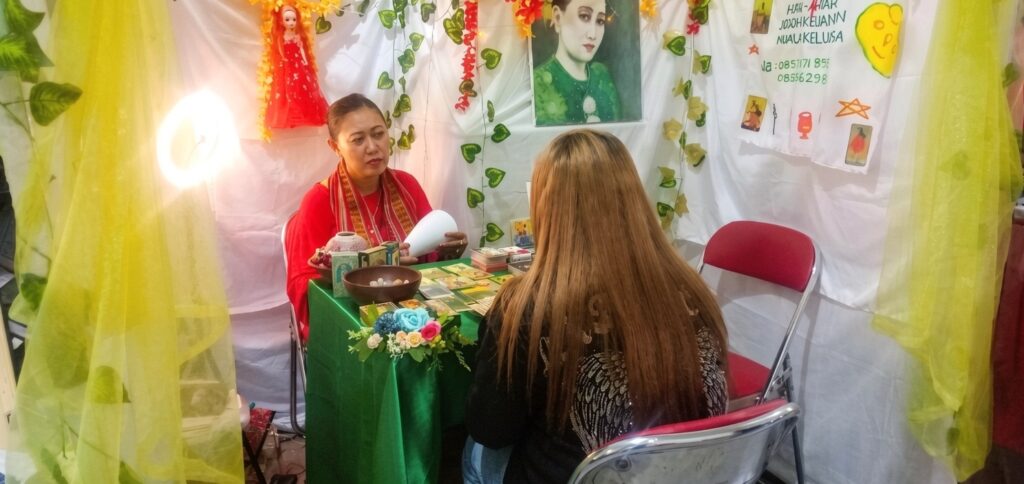 Fortune-teller in red reads cards to a client at a green-clothed table; booth is decorated with flowers and hanging yellow fabric.