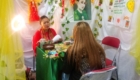 Fortune-teller in red reads cards to a client at a green-clothed table; booth is decorated with flowers and hanging yellow fabric.
